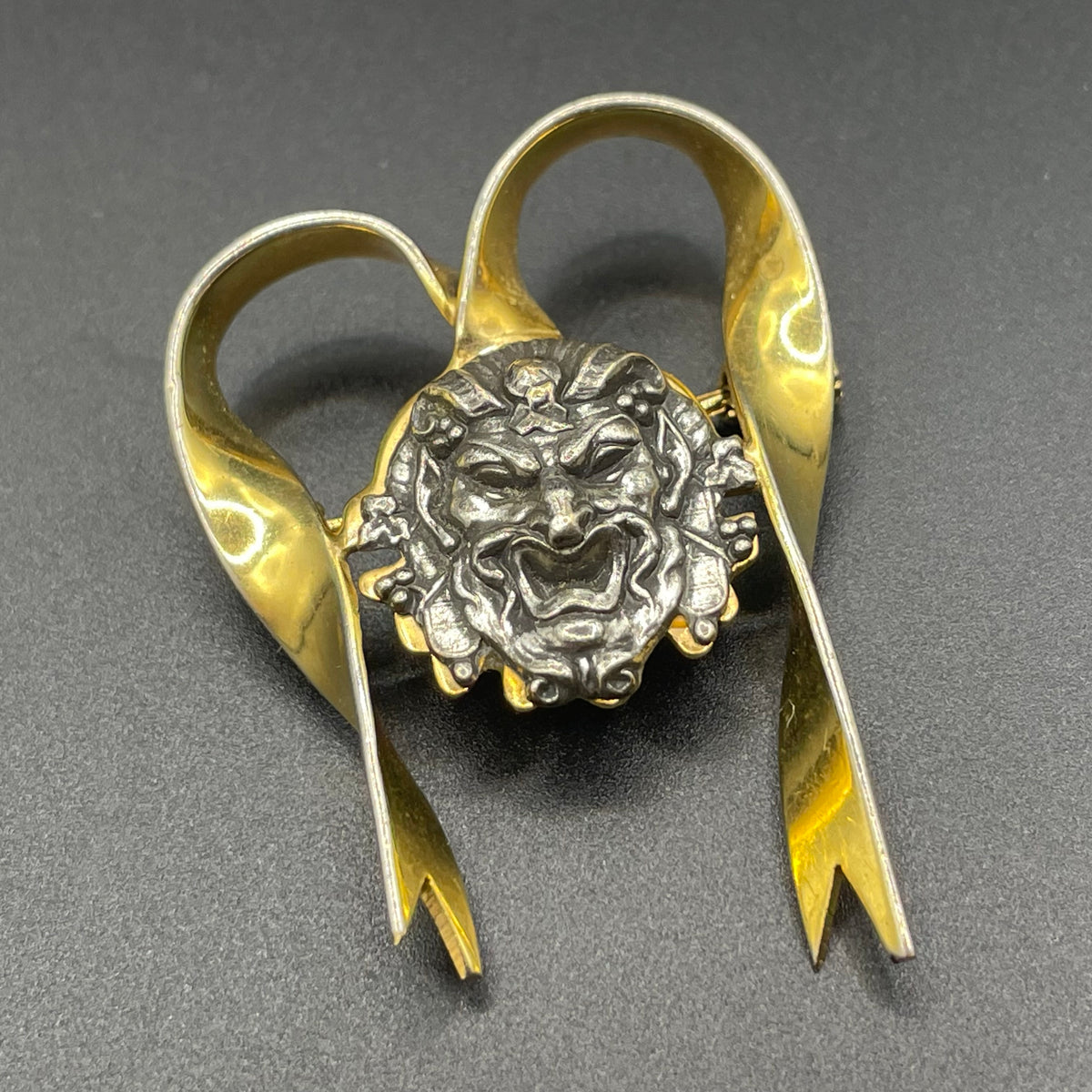 Unusual vintage 'Bacchus' face brooch with silver tone mounted on gold tone ribbon motif, French machine age construction, trombone clasp