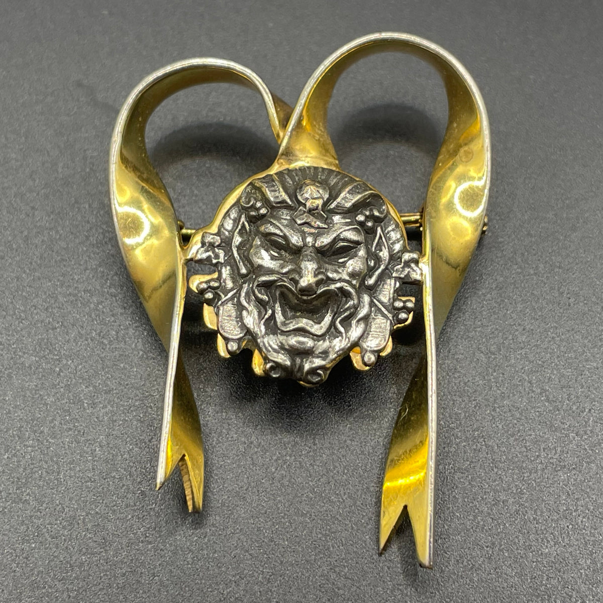 Unusual vintage 'Bacchus' face brooch with silver tone mounted on gold tone ribbon motif, French machine age construction, trombone clasp