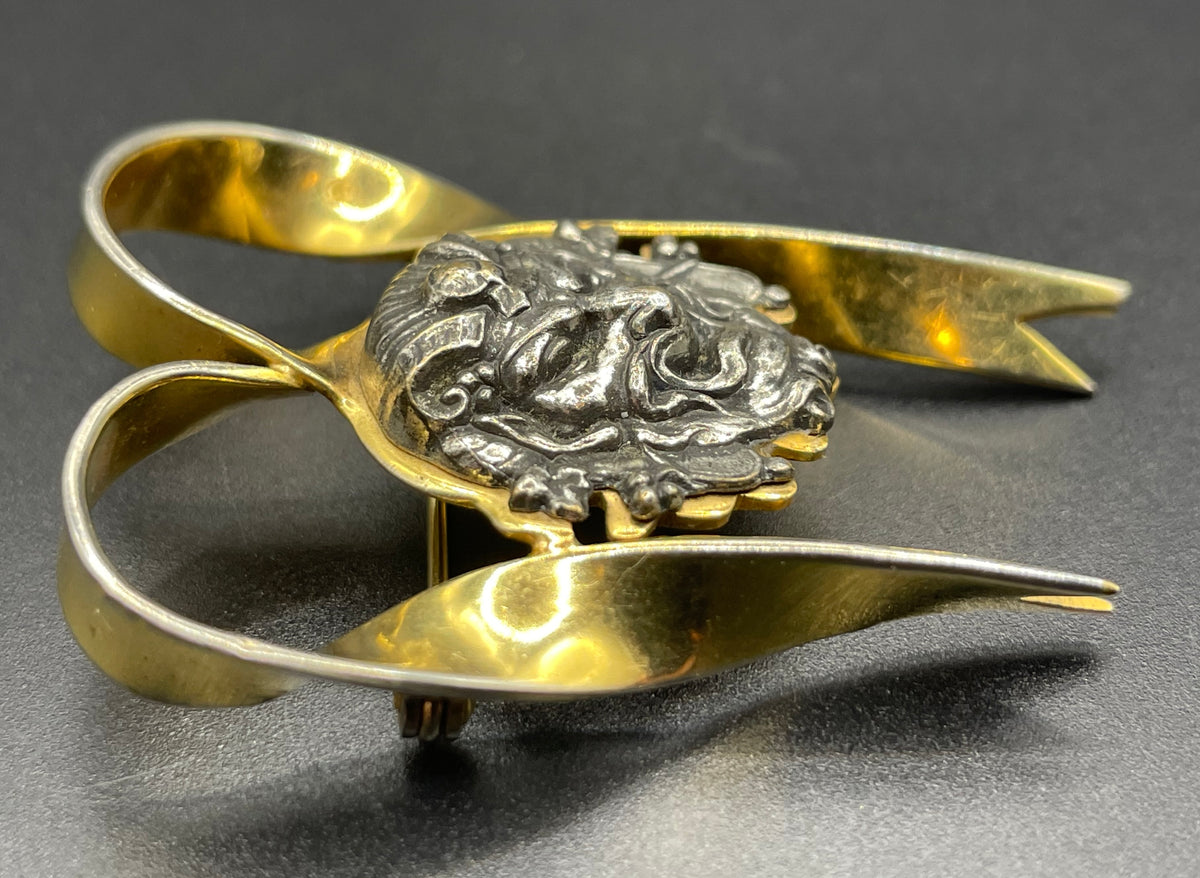 Unusual vintage 'Bacchus' face brooch with silver tone mounted on gold tone ribbon motif, French machine age construction, trombone clasp